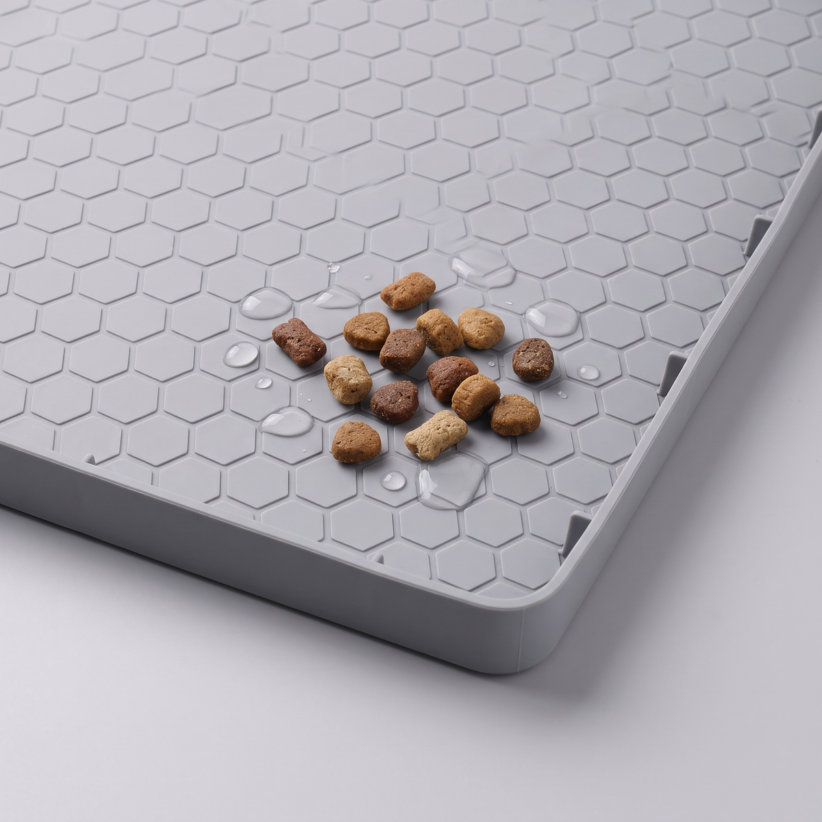 Close-up of the raised edge of a gray feeding mat beside kibble and water droplets